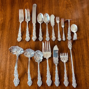 May include: A collection of silver-toned flatware featuring ornate floral and scrollwork handles. The set includes various forks, spoons, knives, ladles, and specialized serving utensils, all displaying intricate embossed detailing on the handles.
