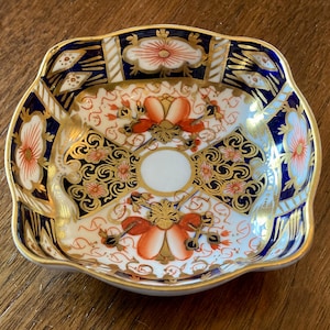 May include: A small, square-shaped dish with a scalloped edge. The dish features a white base with intricate patterns in navy blue, gold, and orange. Floral and geometric designs adorn the surface, with a gold trim around the edge.