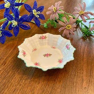 May include: A white porcelain bowl with a scalloped edge and a green trim. The bowl is decorated with a floral pattern of pink roses.