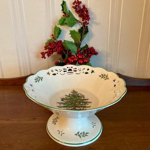 May include: A white ceramic pedestal bowl with a green rim and a Christmas tree design. The bowl has a scalloped edge with cut-out details. Holly berries and leaves are arranged behind the bowl.