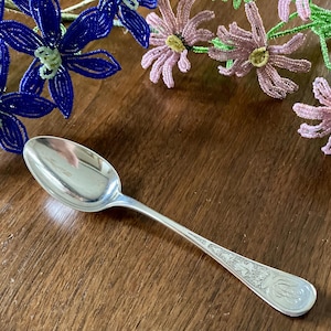 May include: A silver spoon with an ornate handle lies on a wooden surface, with beaded flower decorations in the background. The spoon's bowl reflects the surrounding environment. The handle features detailed engravings.