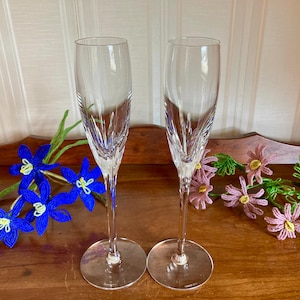 May include: Two clear crystal champagne flutes with etched vertical lines. The flutes sit on a wooden surface, with beaded blue and pink floral decorations. The glasses have a long stem and a round base.