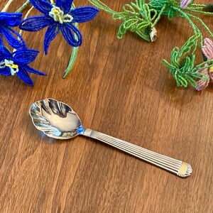 May include: A silver sugar spoon with a gold-colored rim and a decorative, scalloped bowl. The spoon is lying on a wooden surface.