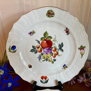 May include: White ceramic plate with a gold rim and a decorative fruit and floral design. The center features a large red apple, purple rose, and various other fruits and berries. The plate has a textured border.