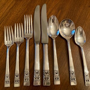 May include: A set of seven silver-toned stainless steel flatware pieces including three forks, two knives, and two spoons. Each utensil features a polished finish with a vertical ribbed handle and a detailed floral embossed design at the base.