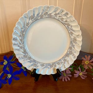 May include: A white porcelain plate with a scalloped edge and a floral pattern in gray and silver. The plate is displayed on a black stand, with beaded flower decorations in the foreground.