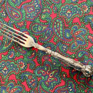 May include: A silver fork with an ornate handle, resting on a red and green paisley patterned fabric.