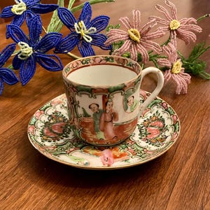 May include: A white porcelain teacup and saucer with a floral and Asian-inspired design. The cup and saucer have a pink, green, and gold pattern.