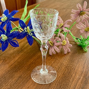 May include: A clear glass shot glass with a cut crystal design. The glass has a tall, slender stem and a round base. The glass is sitting on a wooden table.