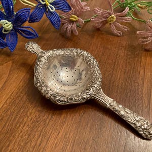 May include: Ornate silver tea strainer with a floral design. The strainer has a round bowl with small holes and a long handle. Decorative beaded flowers in blue and pink are in the background.