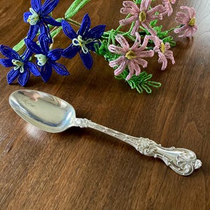 May include: A silver spoon with an ornate handle rests on a wooden surface. The spoon's bowl is reflective, and the handle features detailed floral and scrollwork. Beaded blue and pink flowers are in the background.