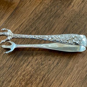 May include: A silver sugar tong featuring claw-shaped ends and a floral embossed handle. The metal surface is engraved with the text "A. LYNX & SON CO" and "STERLING". The design includes intricate botanical details along the handle and bird-like talons.