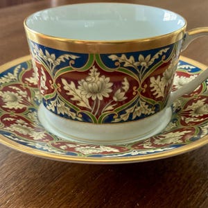 May include: A porcelain teacup and saucer set featuring a repeating pattern of white floral motifs set against deep red and blue backgrounds. The rim of the cup and the edge of the saucer are finished with a thin gold band, and the cup has a handle.