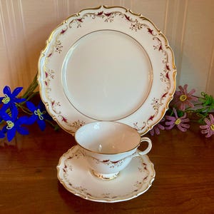 May include: A vintage tea set featuring a large white plate, a teacup, and a saucer. The set has a gold trim with burgundy accents. The teacup and saucer are placed in front of the plate. Decorative blue and purple flowers are in the background.
