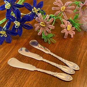 May include: Three silver-toned butter spreaders with rounded blades and ornate, embossed floral detailing on the handles. The metal utensils feature a polished, reflective finish and are arranged diagonally on a wooden surface near beaded flowers.