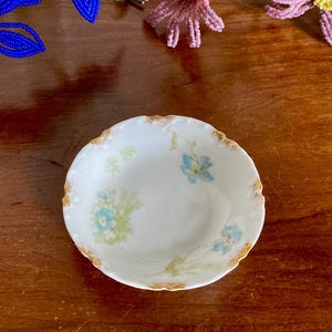 May include: A small, white porcelain dish with a scalloped, gold-trimmed edge. The dish is decorated with delicate blue and green floral patterns. The design includes small blue flowers and green leaves. The dish is likely vintage or antique.