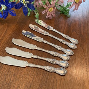 May include: Set of six silver butter knives with ornate handles. The knives have a polished finish and are arranged on a wooden surface.