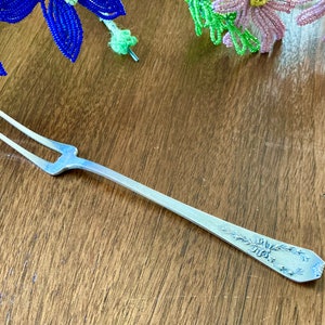 May include: A silver serving fork with a decorative floral design on the handle. The fork has two prongs and is approximately 7 inches long.