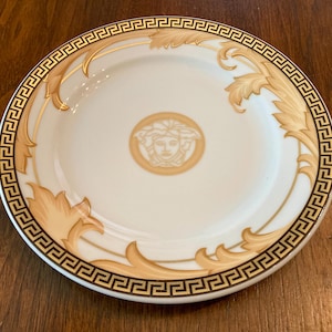 May include: A white ceramic plate with a gold and brown patterned design. The plate features a stylized face in the center and a repeating geometric border.