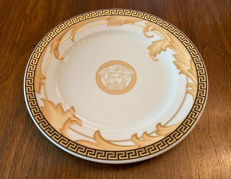 May include: A white ceramic plate with a gold and brown patterned design. The plate features a stylized face in the center and a repeating geometric border.