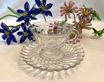 May include: A clear glass teacup and saucer with a swirl design. The cup has a handle and the saucer has a scalloped edge.