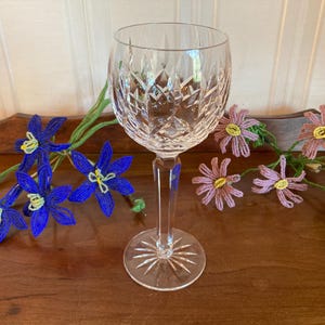 May include: A clear crystal wine glass with a detailed cut pattern. The glass is on a wooden surface, with beaded blue and pink flower garlands. The glass has a starburst design on the base.