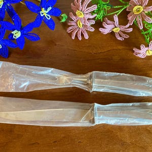 May include: A silver cake server and knife set, still in their original plastic packaging, are displayed on a wooden surface. Decorative beaded flowers in blue, pink, and yellow are arranged around the utensils.