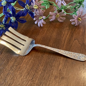 May include: Silver serving fork with a floral pattern on the handle. The fork has four tines and is resting on a dark wooden surface. Decorative beaded flowers in blue and pink are in the background.