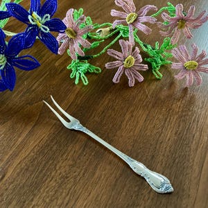 May include: A silver-toned metal cocktail fork featuring a two-pronged head and an ornate, embossed floral pattern along the handle. The fork is positioned diagonally, accompanied by a decorative arrangement of blue and pink beaded flowers.