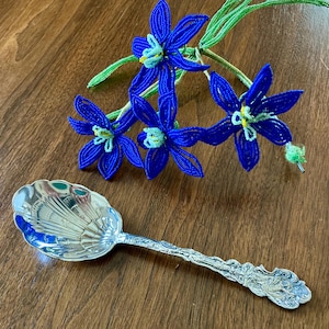 May include: A silver serving spoon with a shell-shaped bowl and ornate handle, next to a bouquet of blue and green beaded flowers.