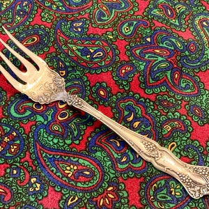 May include: A silver fork with an ornate design featuring grapes and leaves. The fork is on a red and green paisley patterned fabric.