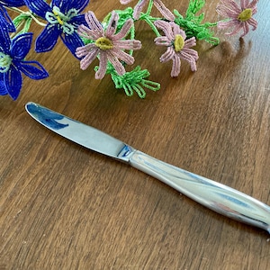 May include: A silver butter knife with a curved handle rests on a wooden surface. Beaded blue and pink floral decorations are in the background. The knife is approximately 8 inches long.