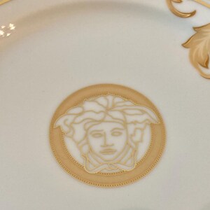 May include: A white ceramic plate with a gold and black border. The center of the plate features a gold and white image of a Medusa head.
