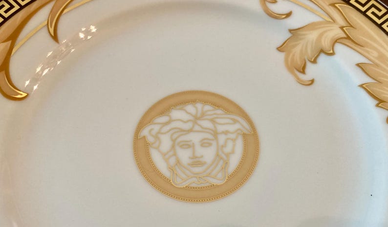 May include: A white ceramic plate with a gold and black border. The center of the plate features a gold and white image of a Medusa head.
