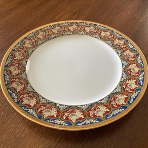 May include: A round ceramic dinner plate with a plain white center and a wide decorative border. The border features a repeating pattern of cream-colored floral motifs set against a red and blue background, finished with a thin gold rim.