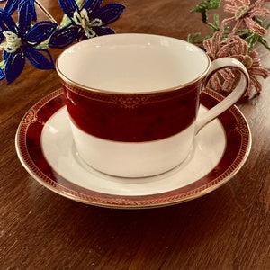 May include: A red and gold teacup and saucer set with a delicate floral design. The cup and saucer are both trimmed with a gold band.