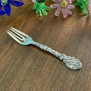 May include: A silver fork with an ornate handle, resting on a wooden surface. The fork has three tines and a detailed floral design on the handle. Decorative beaded flowers in blue, green, and pink are in the background.