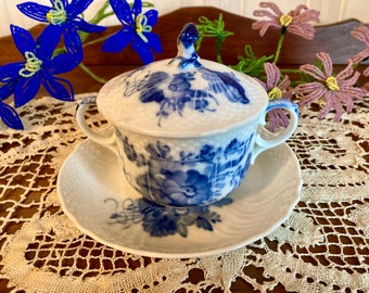 Op de afbeelding: Een witte porseleinen roomkan met een blauw bloemenpatroon en een deksel. De roomkan staat op een wit schoteltje met een blauw bloemenpatroon. De roomkan en het schoteltje staan op een wit kantkleed.