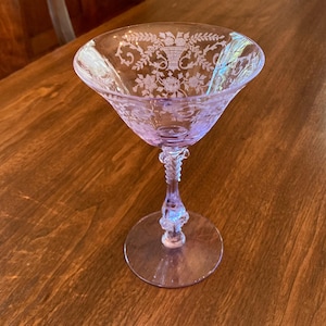 May include: A light purple glass cocktail coupe featuring an etched floral pattern around the rim. The stem has a decorative molded design leading down to a clear circular base. The glass has a wide, shallow bowl and a translucent finish.