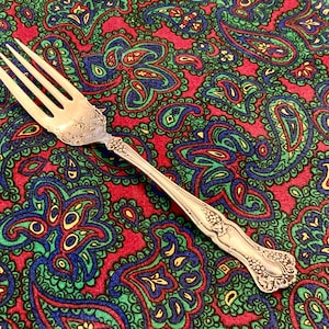 May include: A silver fork with an ornate design on a red, green, and blue paisley patterned fabric.
