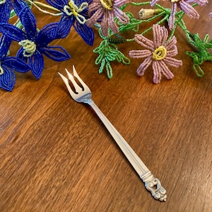 May include: A silver fork with three tines, resting on a wooden surface. The handle has a decorative design at the end. Beaded blue and purple flowers are in the background, adding a touch of color and artistry to the scene.