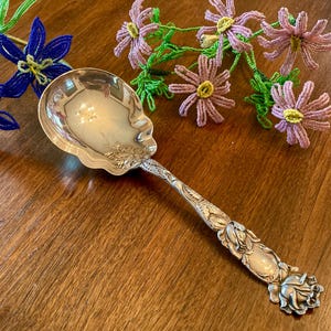 May include: A silver serving spoon with ornate floral detailing. The spoon has a scalloped bowl and a handle adorned with roses and leaves. Decorative beaded flowers in blue and purple are in the background.