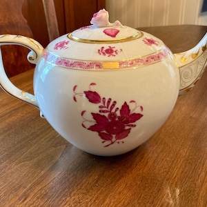 May include: White porcelain teapot with a floral design in shades of pink and gold accents. The teapot has a rounded body, a curved spout, and a handle. The lid is topped with a pink flower. The teapot is sitting on a wooden surface.