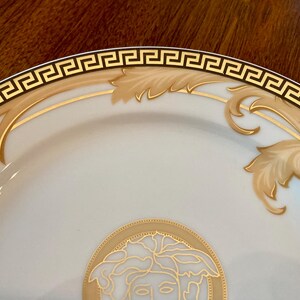 May include: A white ceramic plate with a gold and black Greek key border. The plate features a gold floral design and a gold image of a Medusa head in the center.