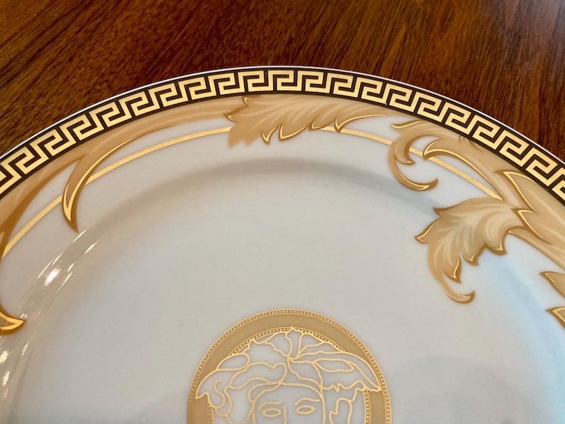 May include: A white ceramic plate with a gold and black Greek key border. The plate features a gold floral design and a gold image of a Medusa head in the center.