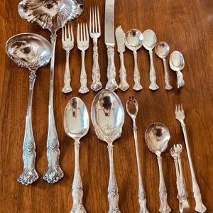 May include: A collection of silver-toned metal flatware and serving utensils featuring ornate floral and vine embossed handles. The set includes two large ladles, several forks, spoons, a knife, a butter spreader, sugar tongs, and a long-handled fork.