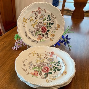May include: Set of four white ceramic dinner plates with a floral pattern in shades of pink, yellow, green, and brown. The plates have a scalloped edge.