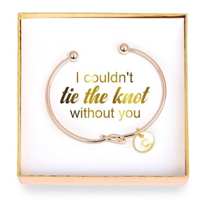 Könnte beinhalten: Ein goldfarbenes Armband mit einem Knoten-Anhänger und einem kleinen Anhänger mit dem Buchstaben "C". Das Armband befindet sich in einer weißen Schachtel mit goldfarbenem Rand. Die Schachtel trägt den Text "I couldn't tie the knot without you".