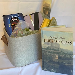 Può includere: Un cesto di stoffa grigia pieno di libri e oggetti decorativi. I libri includono "Throne of Glass" di Sarah J. Maas. Il cesto ha manici in pelle marrone. Altri oggetti includono cancelleria e una stampa arrotolata.
