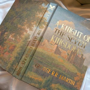 May include: Hardcover book titled "A Knight of the Seven Kingdoms" by George R.R. Martin. The book cover features a landscape painting with gold lettering. The spine also has gold lettering. The book is open.